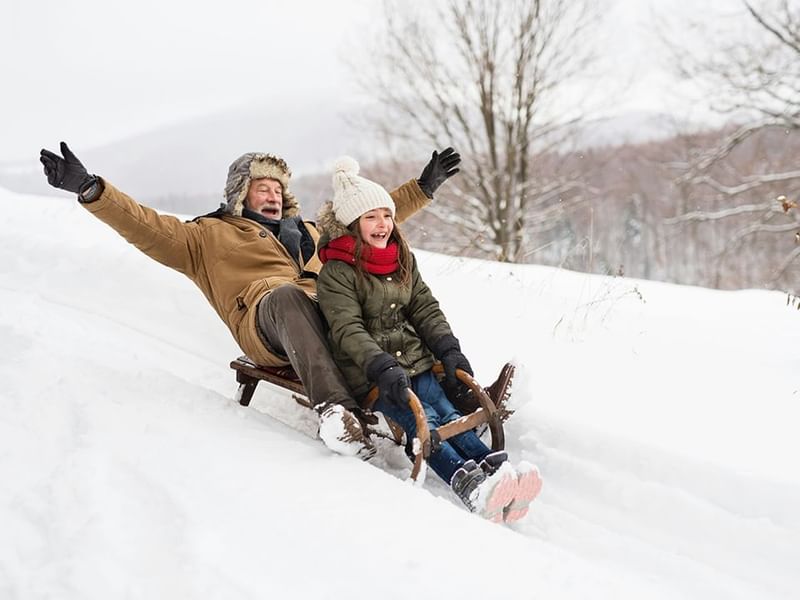 Grandfather and little girl sledding on a wooden sled down snowy hill with arms raised at Mountain View Grand Resort & Spa
