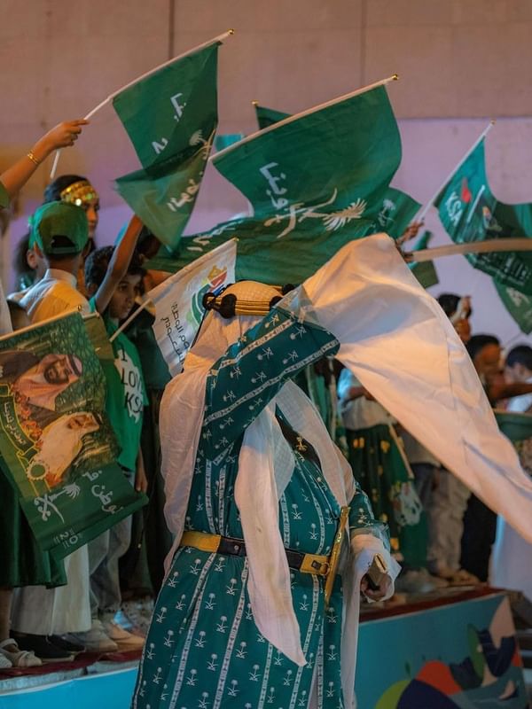 Saudi man in traditional dress waves a white flag while children hold green KSA flags at Saja Warwick Madinah