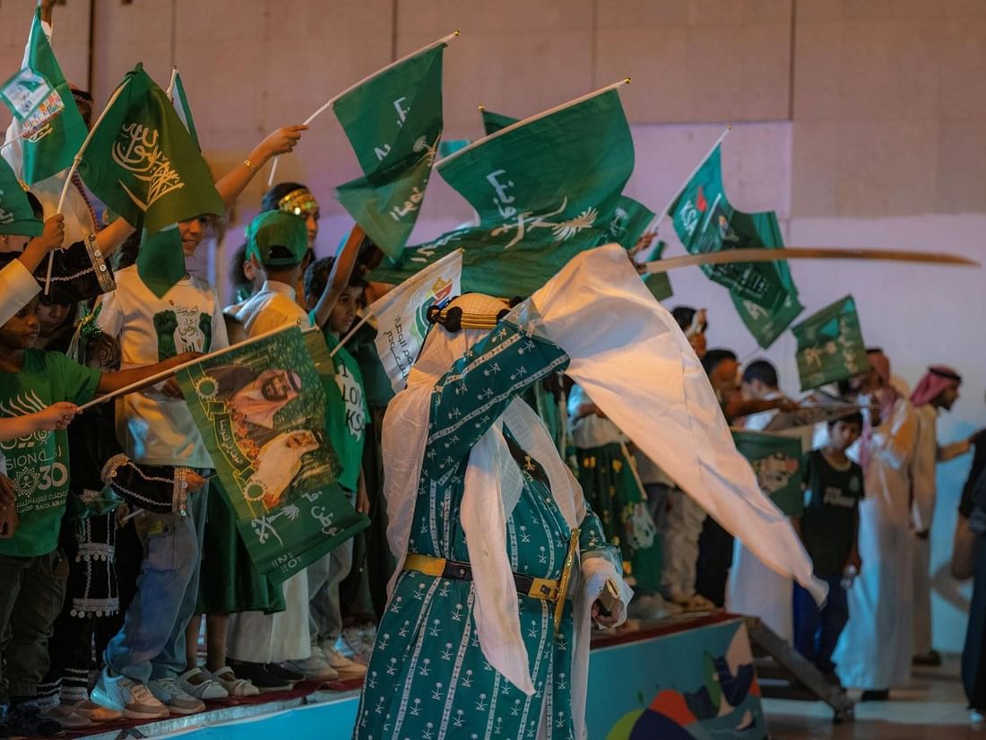 Saudi man in traditional dress waves a white flag while children hold green KSA flags at Saja Warwick Madinah