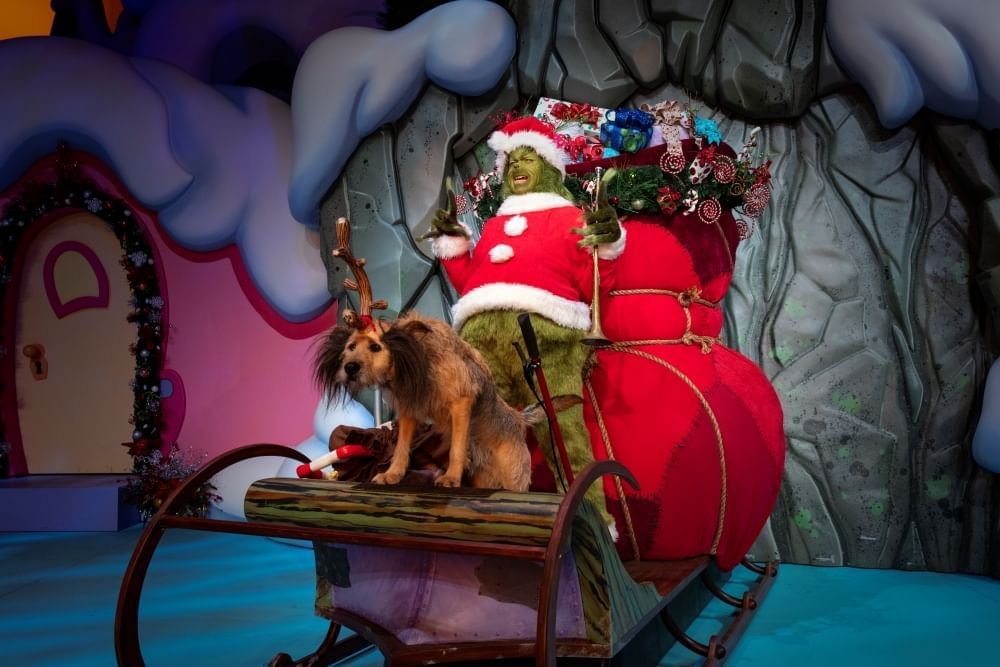 The Grinch in his Santa Suit and a brown scruffy dog ride a sleigh onstage with a huge red sack full of Christmas gifts.