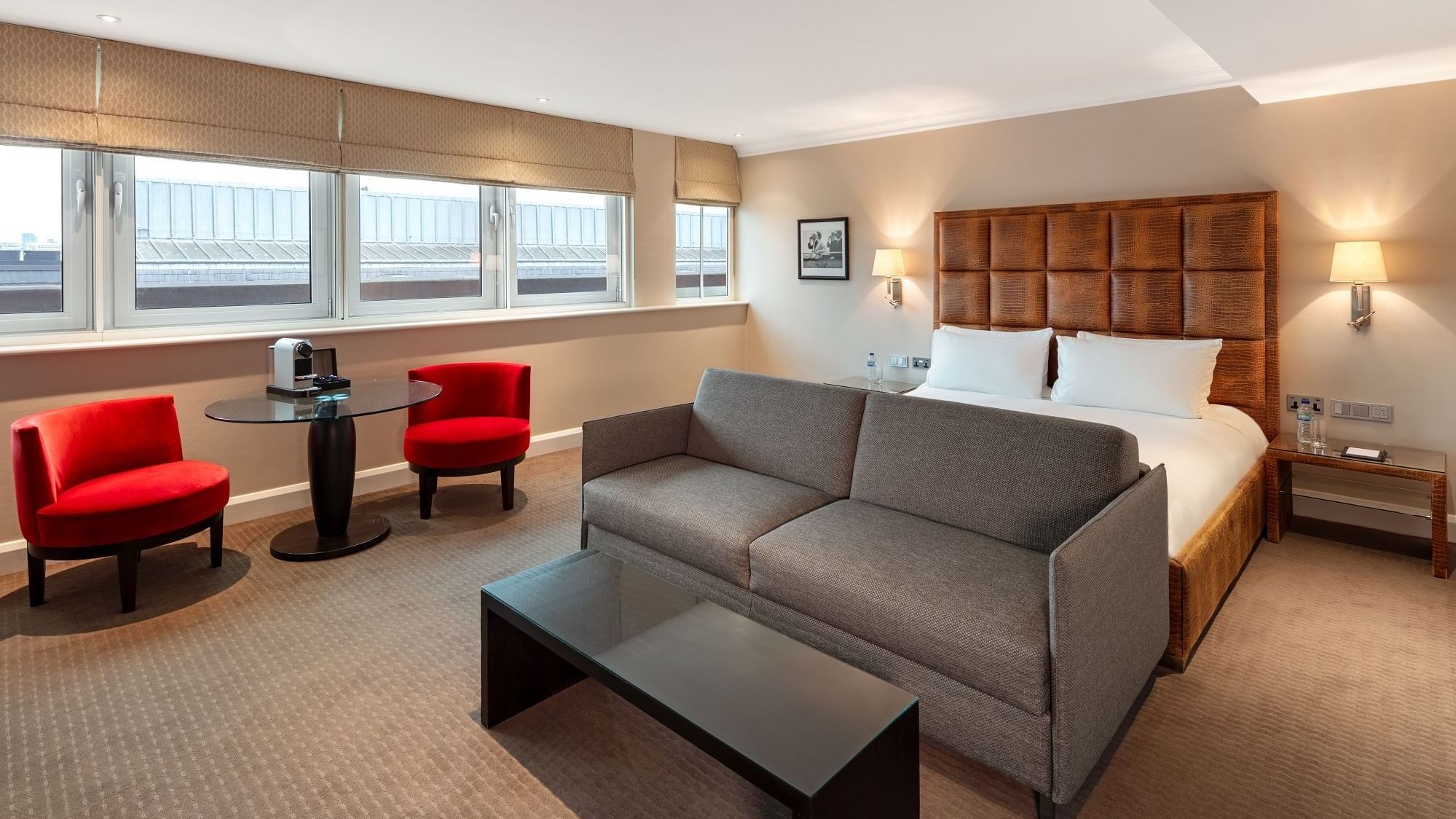 Modern May Fair Premium Deluxe bedroom with king bed, gray sofa, coffee table, and red chairs at The May Fair Hotel