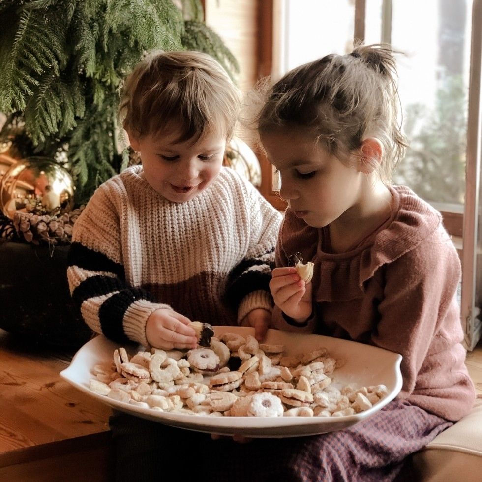 Two children eating powdered sugar cookies from a plate beside a Christmas tree at Falkensteiner Hotels & Residences