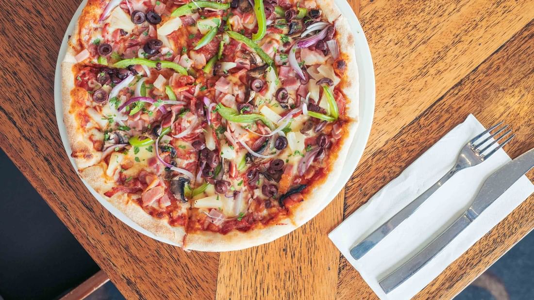 Pizza on wooden table with fork and knife on napkin, ready to be eaten.