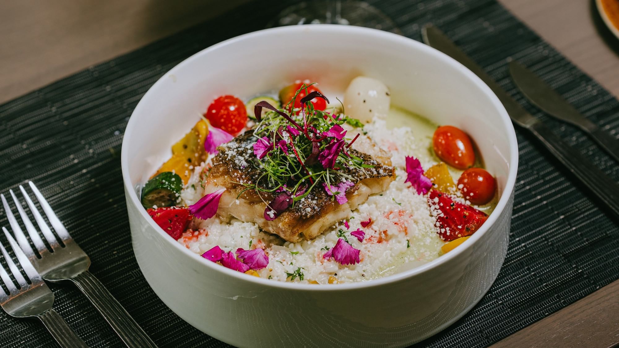 Bowl with fish, rice, tomatoes, and garnish, with cutlery on a placemat.