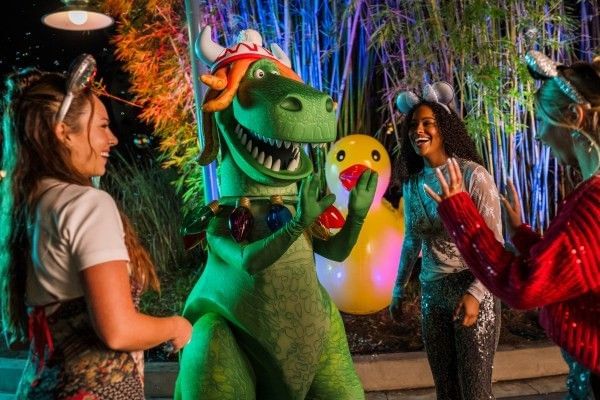 Several adult womenin Disney attire dance around a large T-Rex character wearing a string of Christmas lights and a Viking hat.