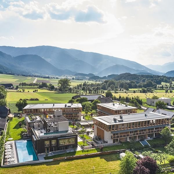 Luftaufnahme eines modernen Resorts mit Schwimmbad und umliegenden Feldern, vor einer Bergkulisse.