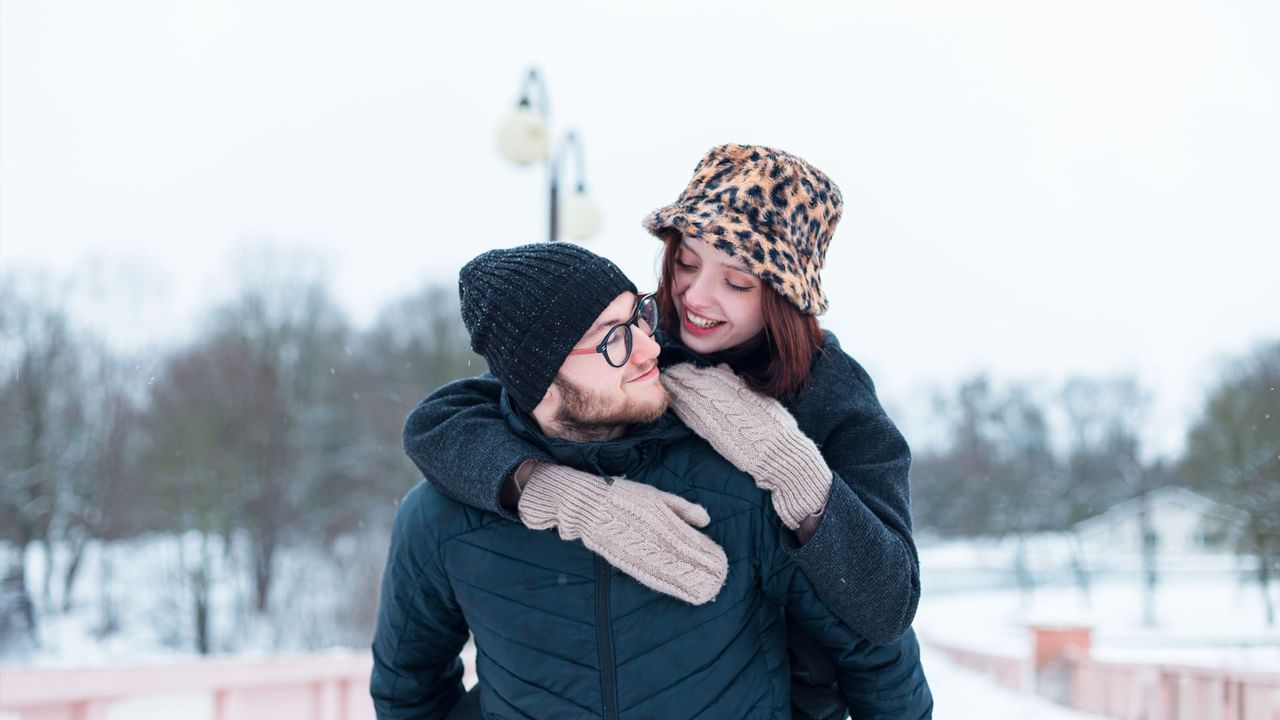 A happy couple enjoying a winter day outdoors, dressed in warm clothing with snow falling around them.