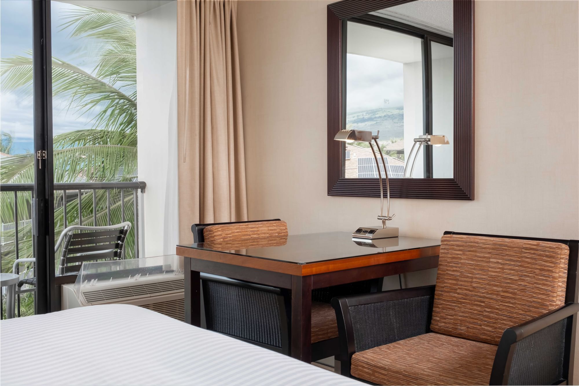 A hotel room with a desk, chair, lamp, and mirror, and a balcony with a view.