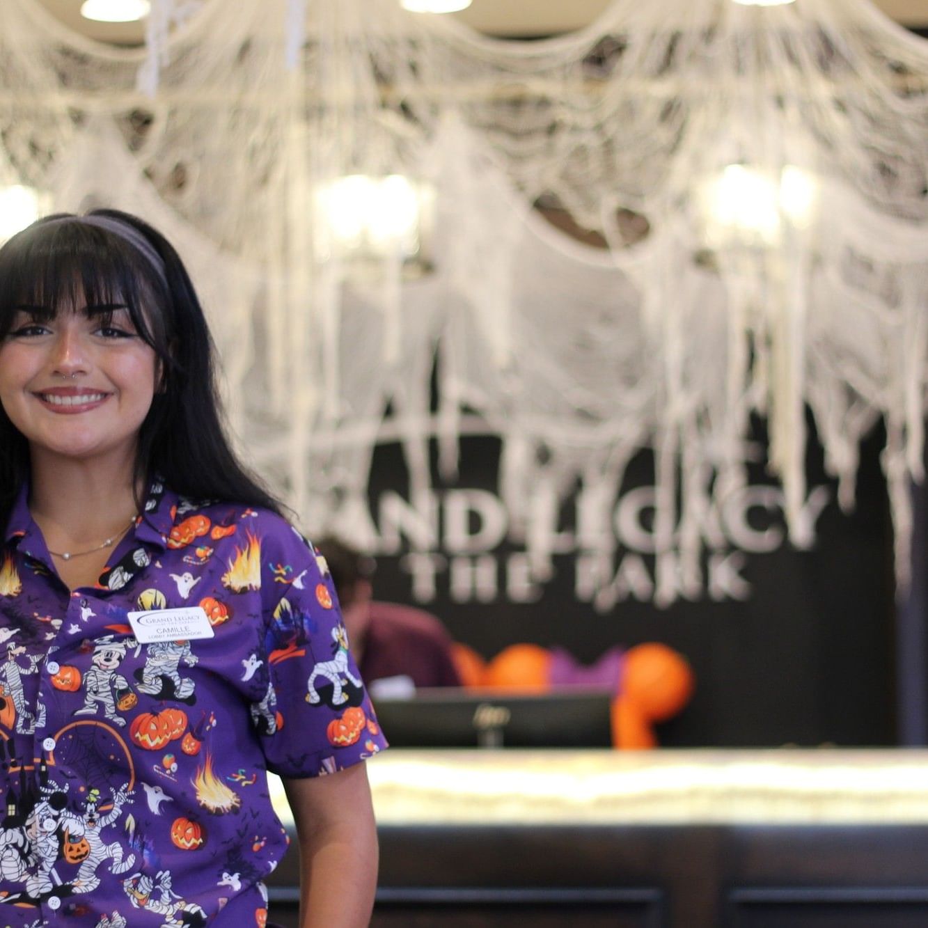 Smiling hotel team member at Grand Legacy At The Park in Anaheim wearing a festive Halloween shirt, standing in front of the decorated hotel lobby with spiderwebs and seasonal décor near Disneyland Resort®