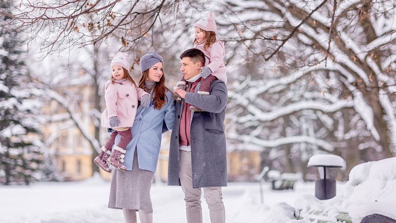A family dressed in winter clothing enjoying a snowy park walk, surrounded by snow-covered trees.