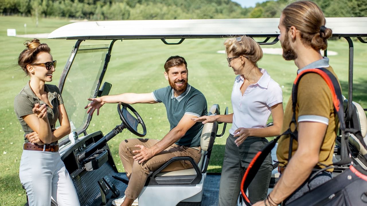 Group of four people conversing next to a golf cart on a green field.