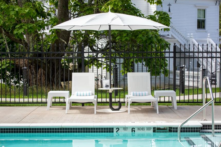 Two white lounge chairs under an umbrella, beside a table with drinks, next to the pool at The Roxbury