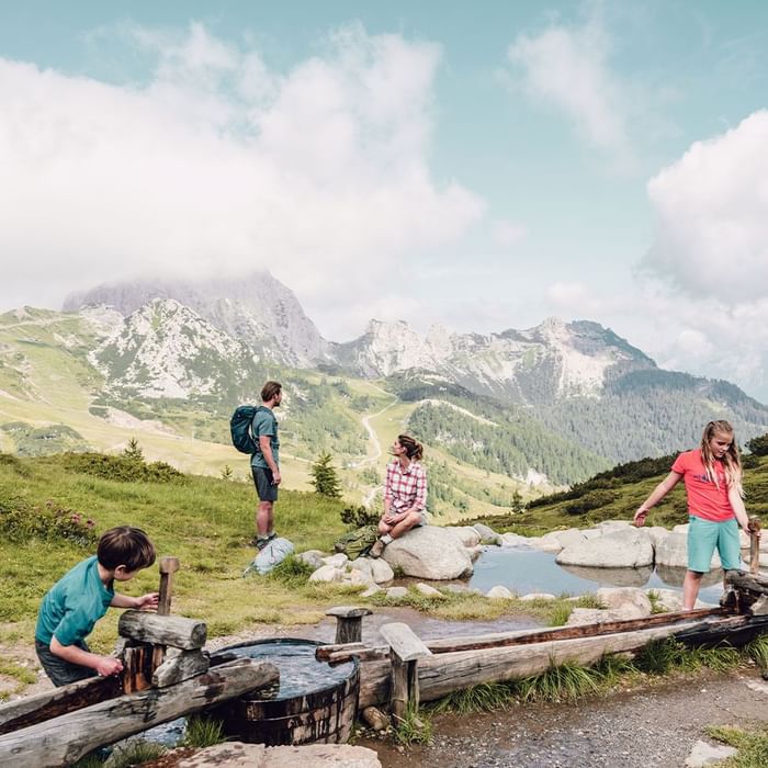 Kinder spielen an einem Wasserrad im Gebirge, während Erwachsene sie beobachten.