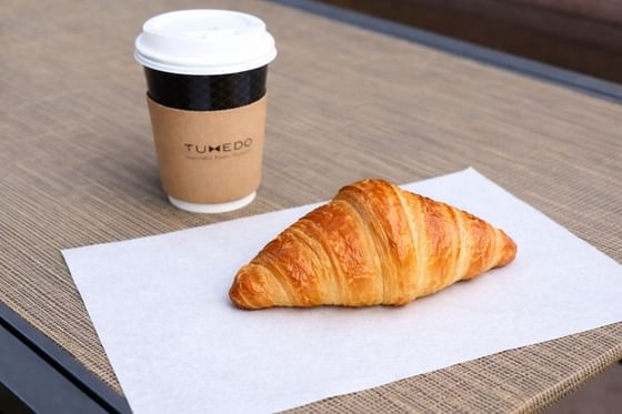 Fresh golden croissant on white paper next to a Tuxedo takeaway coffee cup on a table at Carlton Hotel Singapore