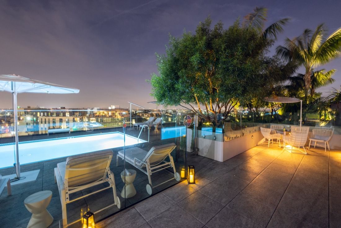 A nighttime view of a rooftop pool area with white furniture and city lights in the background.