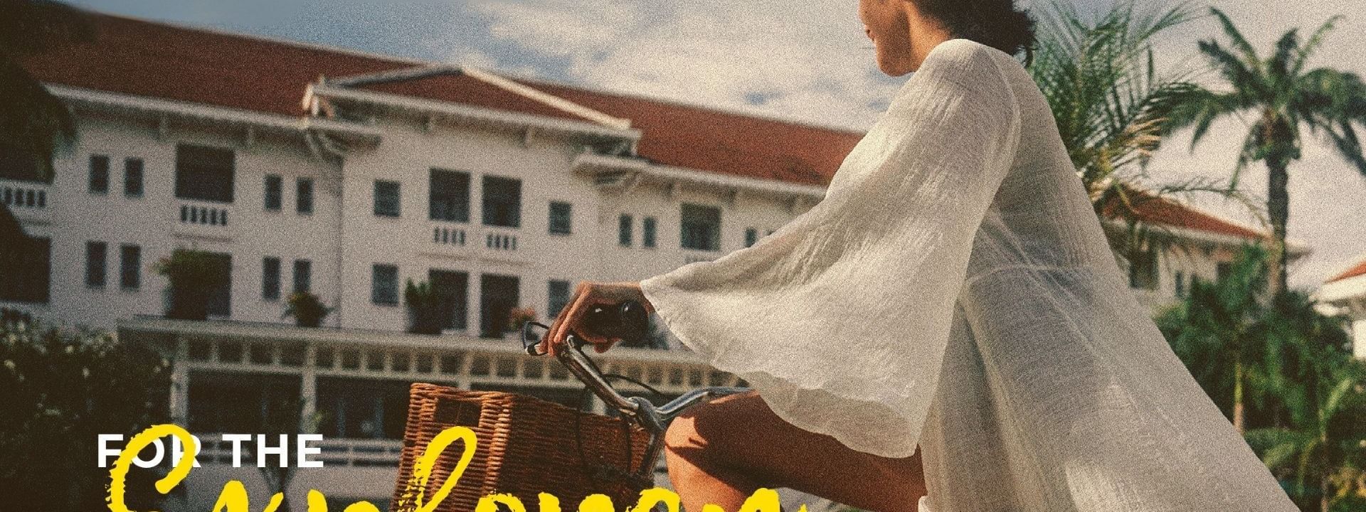 Woman in white robe rides bicycle with basket past resort building with red roof and palm trees.
