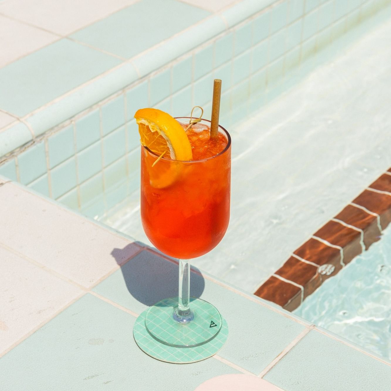 Orange cocktail with ice, orange slice, and cinnamon stick on poolside tiles for Sunday Spritz event