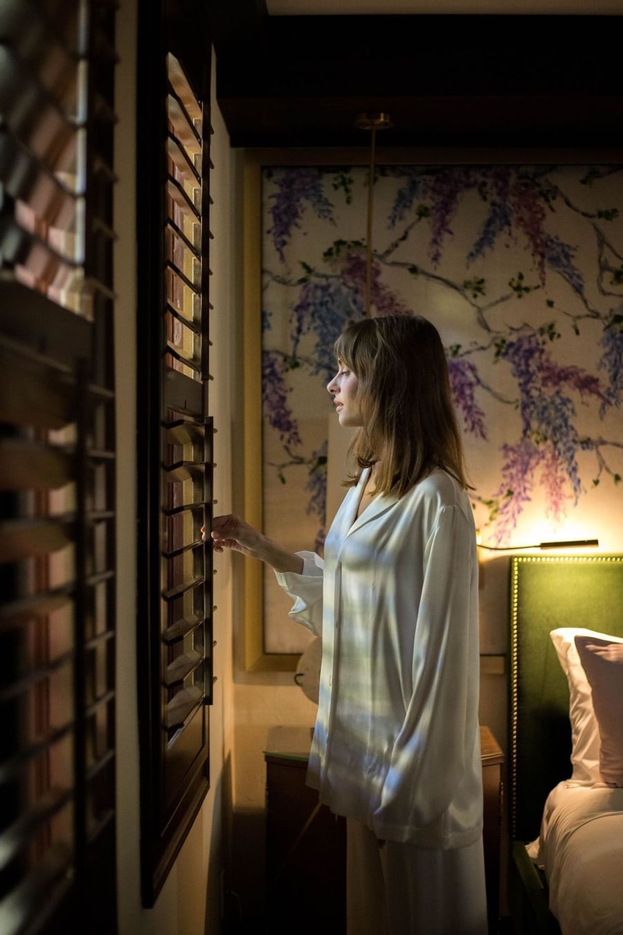 Woman in silk pajamas standing by open wooden shutters near bed with floral artwork at The Brazilian Court