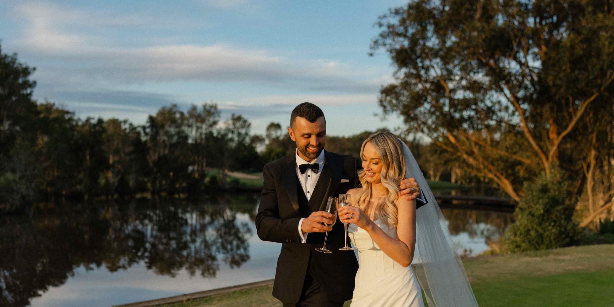 Bride and groom holding champagne glasses by a lake near Mercure Kooindah Waters