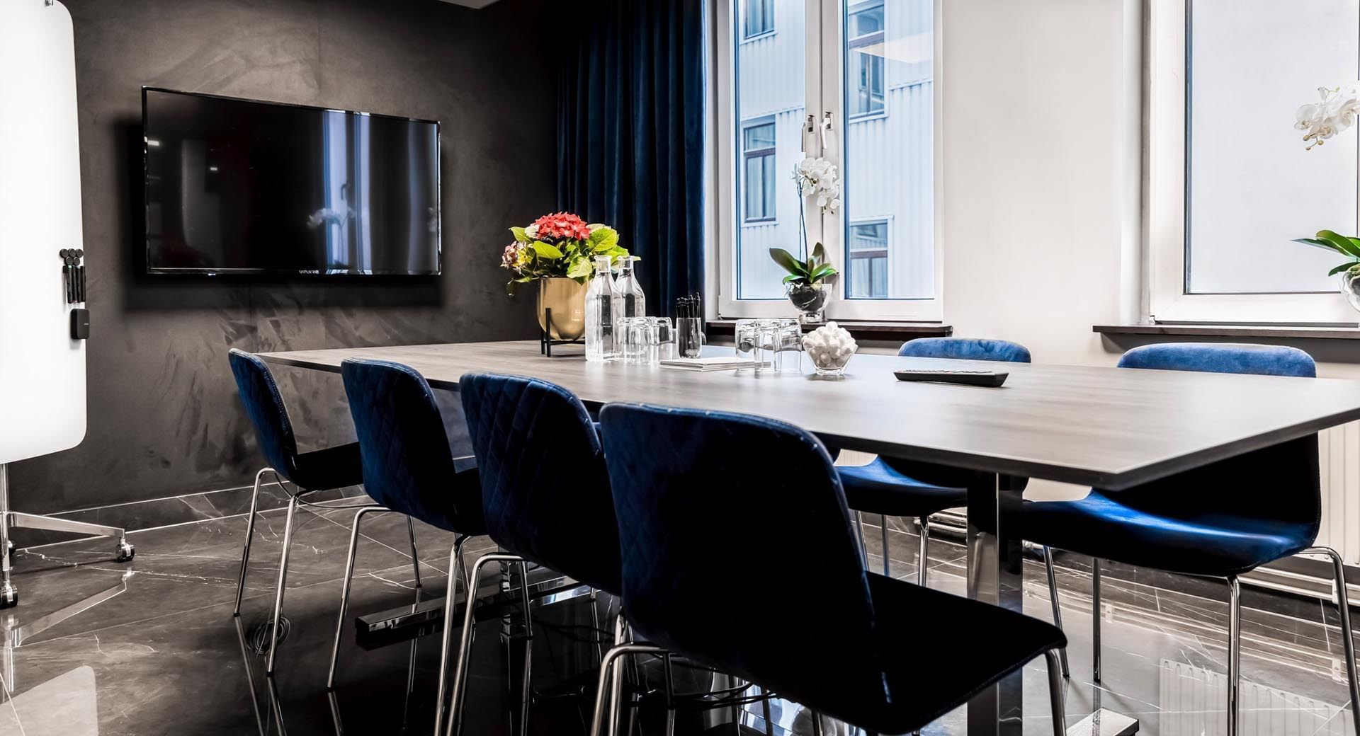 Modern conference room with a large table, blue chairs, TV, flowers, and glass windows.