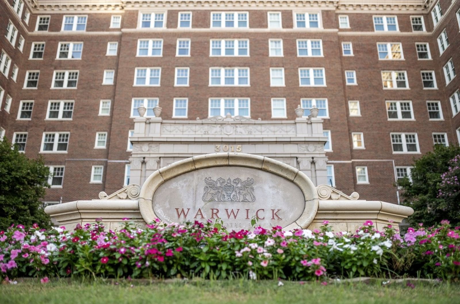 Warwick Hotel logo on a stone sign by pink flowers under the brick facade at Warwick Melrose Dallas