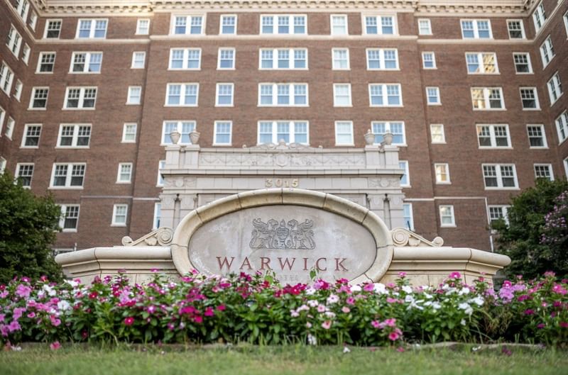 Warwick Hotel logo on a stone sign by pink flowers under the brick facade at Warwick Melrose Dallas