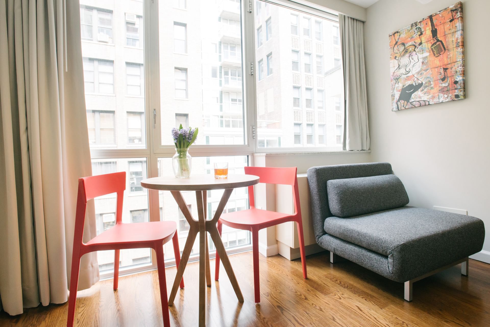 A small seating area with a table, two red chairs, a gray sofa, and a painting on the wall.