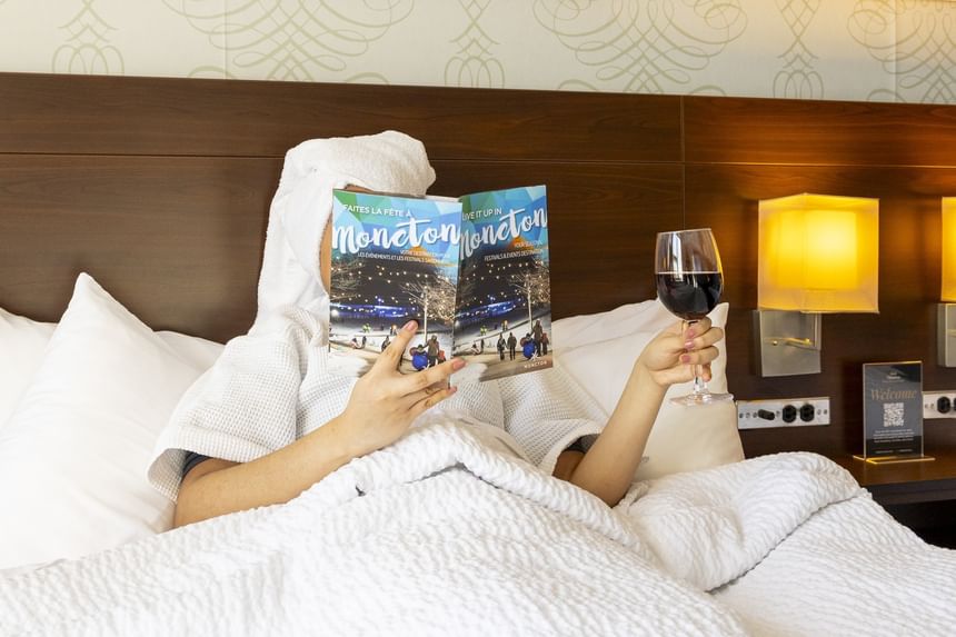 Woman in a white bathrobe relaxing in bed with a glass of red wine while reading a magazine at Rodd Moncton