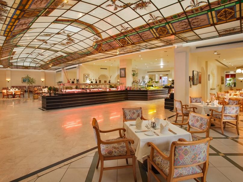 A large, elegant dining room with tables, chairs, and a stained glass ceiling, illuminated by chandeliers.