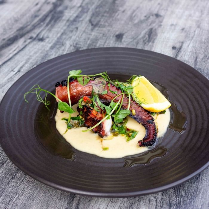 Plate with grilled octopus, creamy sauce, lemon wedge, and green garnish on a wooden surface.