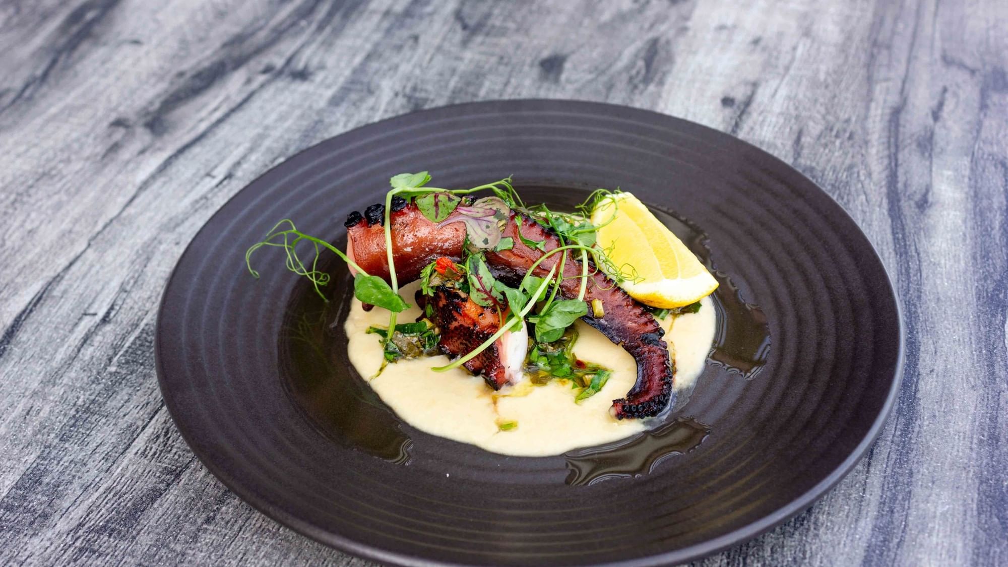 Plate with grilled octopus, creamy sauce, lemon wedge, and green garnish on a wooden surface.