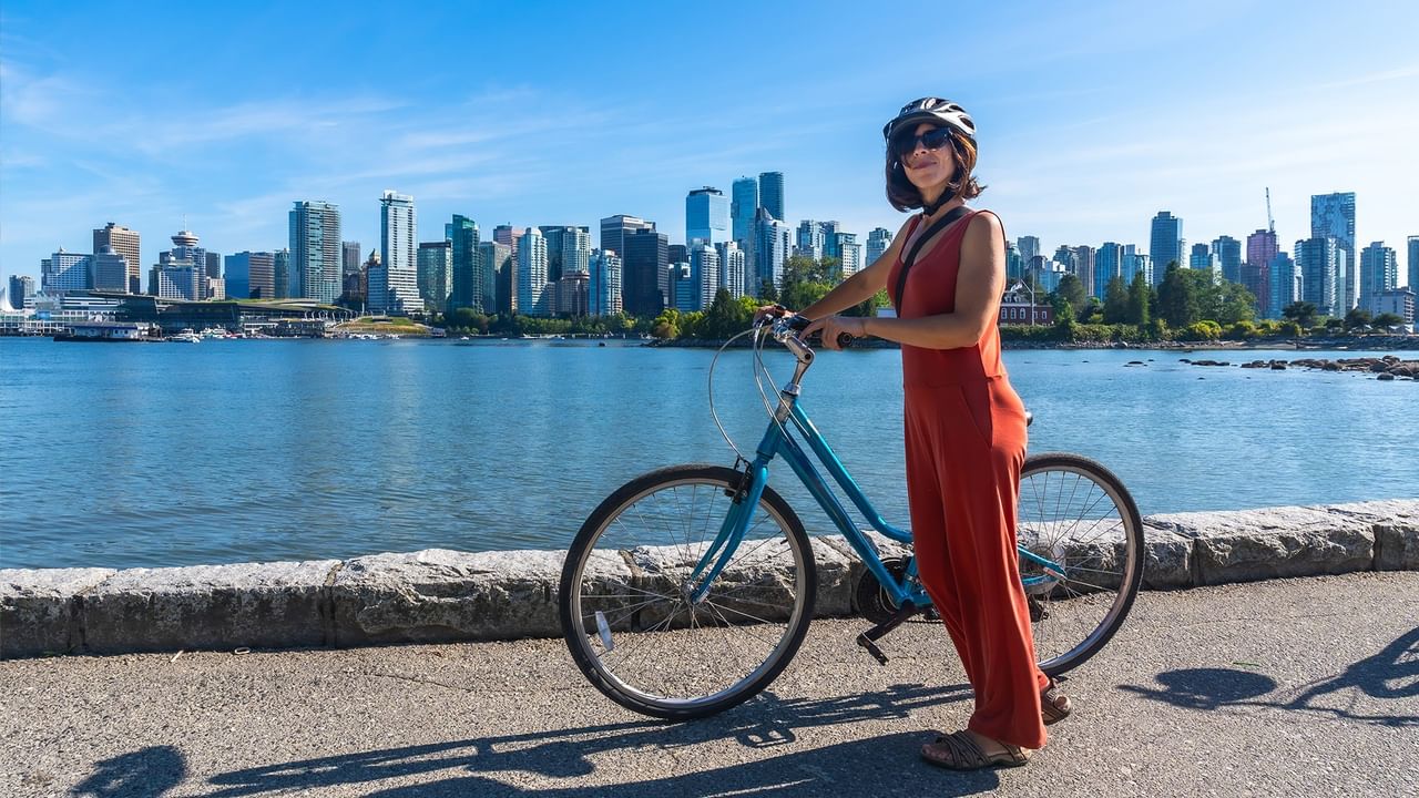 Smiling woman standing by blue bicycle with Vancouver city skyline background.