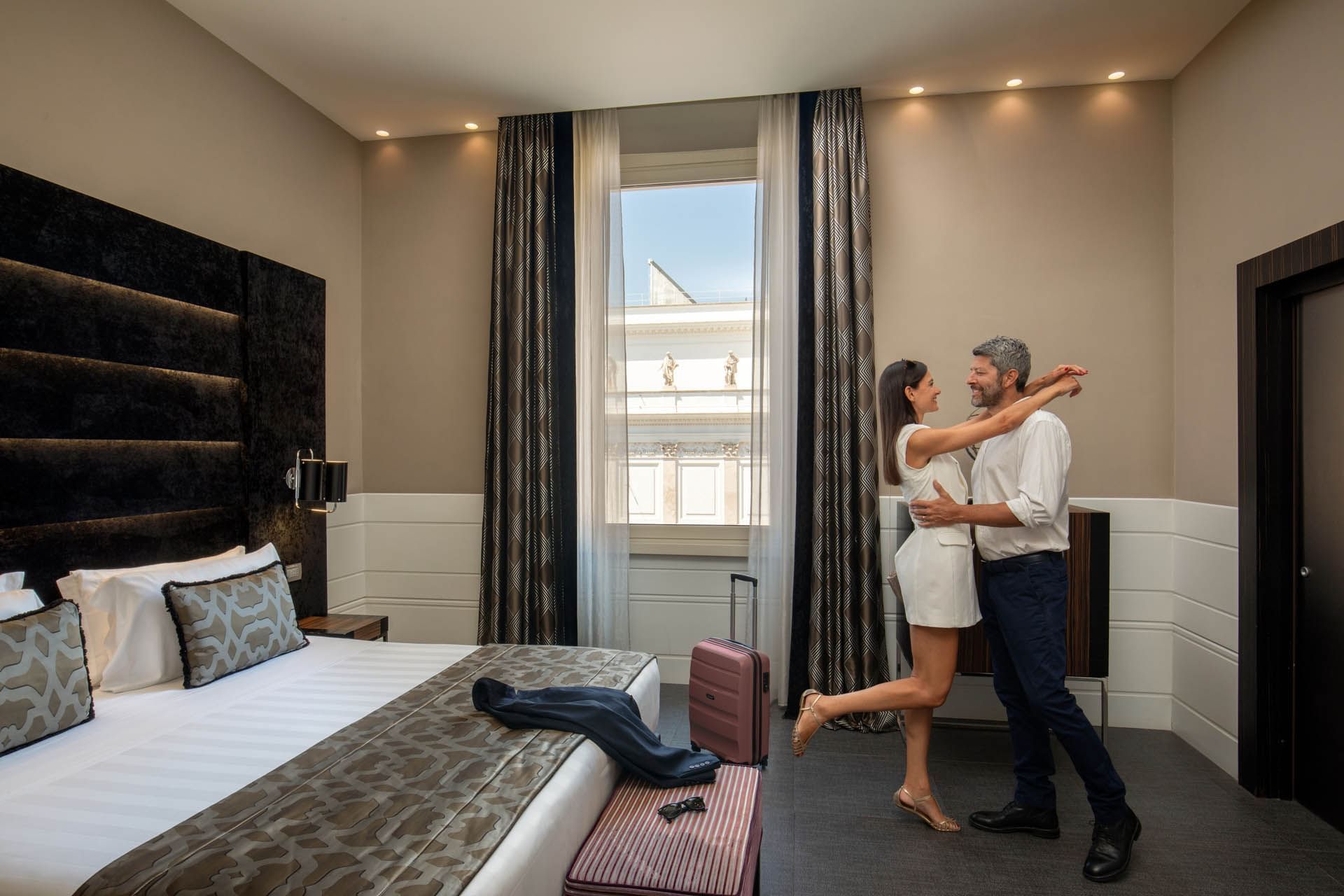 Couple dancing joyfully by the bed with large windows in Superior Room at The Glam Hotel