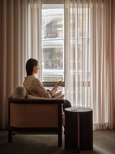 Woman enjoying a drink by the window in a hotel room for The Suite Life offer.