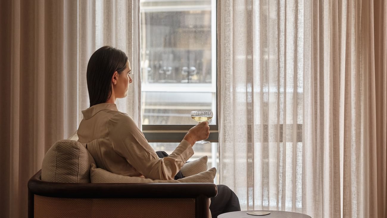 Woman enjoying a drink by the window in a hotel room for The Suite Life offer.