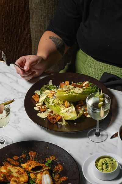 Hand holding a fork over a romaine lettuce salad with croutons, drizzled dressing, & a cocktail with olives at Cypress & Oak