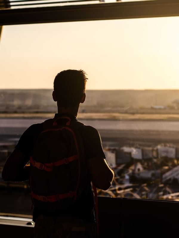 A traveler with a backpack gazes out an airport window at a golden sunset over the runway near Oceania Hotels