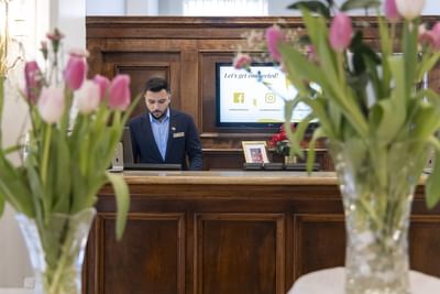 Receptionist standing by a wooden counter behind a vase of pink tulips under soft light at Rodd Charlottetown Hotel