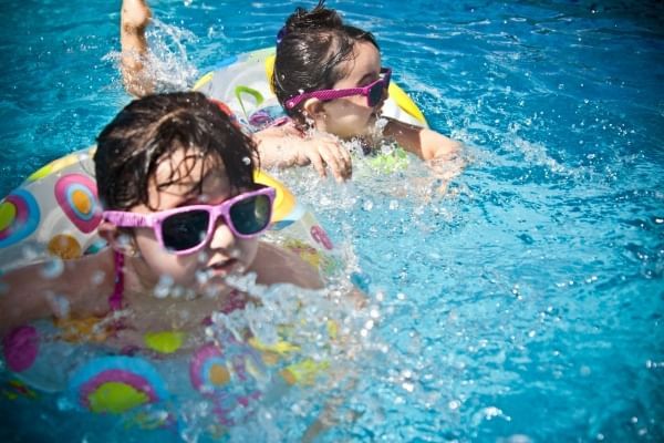 Two kids wearing pink sunglasses in colorful inner tubes splash in a sparkling blue pool. 