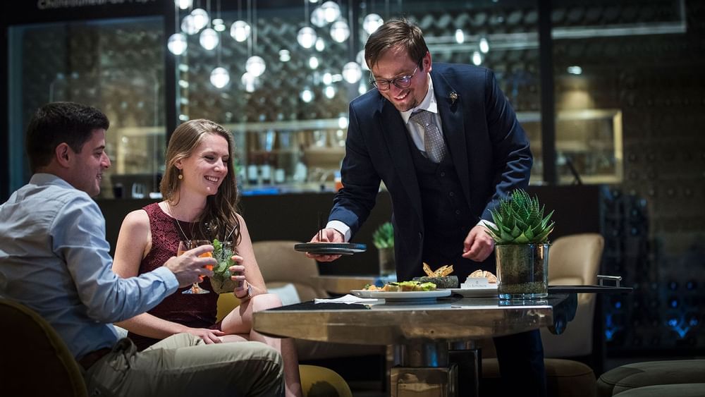 Un serveur en costume présente des plats à un couple souriant dans un restaurant élégant.