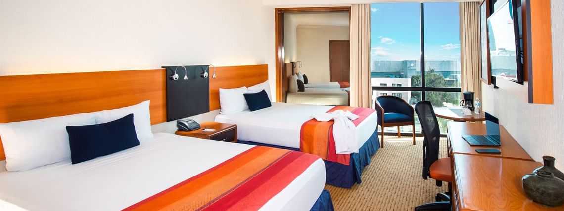 Suite with two beds featuring a desk & an ocean view in the distance, at the Guadalajara Plaza Hoteles & Suites