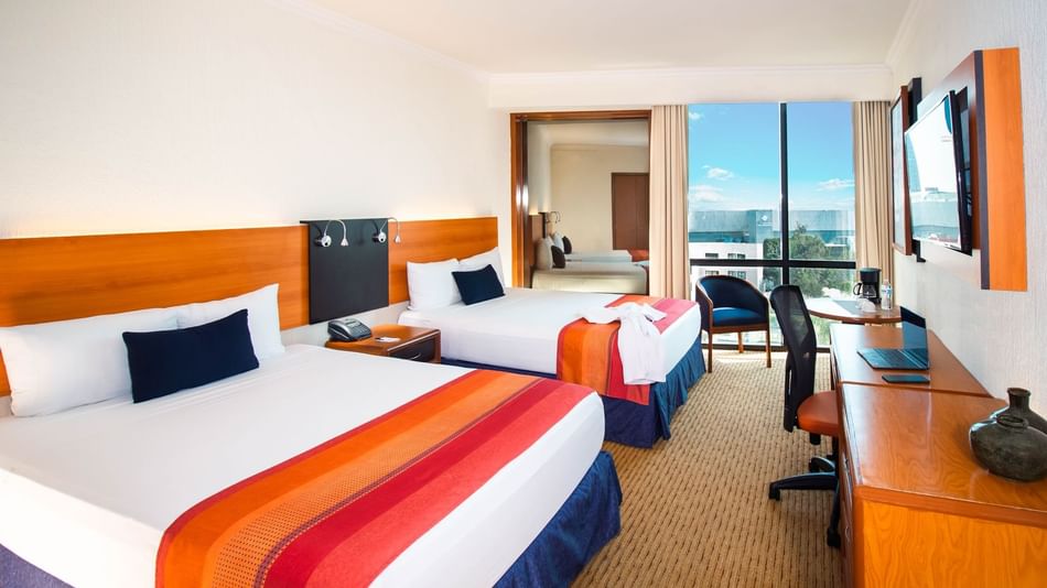 Suite with two beds featuring a desk & an ocean view in the distance, at the Guadalajara Plaza Hoteles & Suites