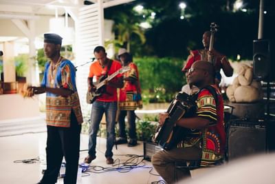Live musicians in colorful dashiki attire playing guitar, accordion, and bass under lights at Hotel Montana Haiti