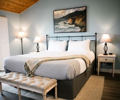 A bedroom with a bed, two nightstands, a bench, and a large painting of a coastal landscape.