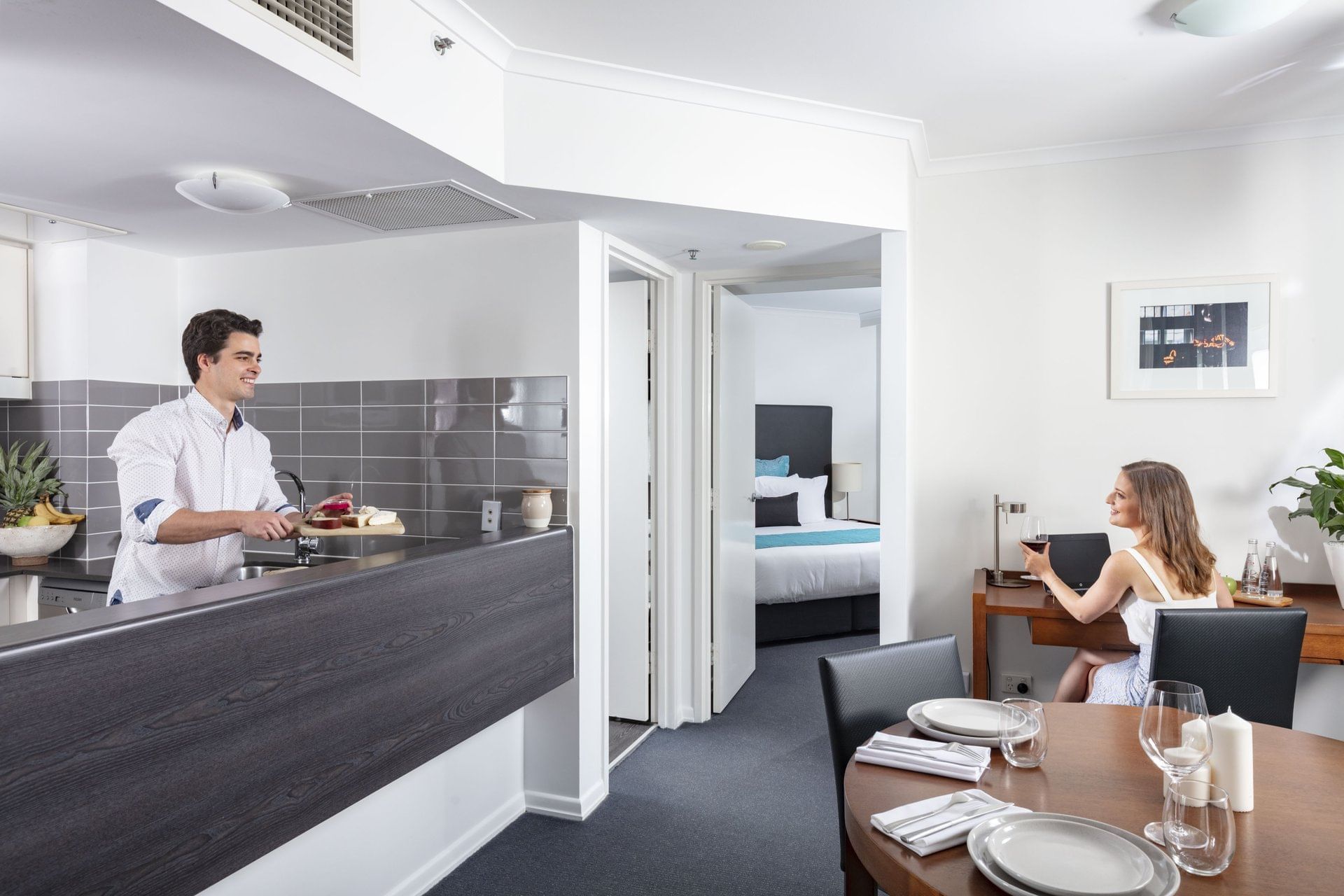 Spacious hotel apartment with a man cooking in the kitchen and a woman working at a desk at The Sebel Brisbane
