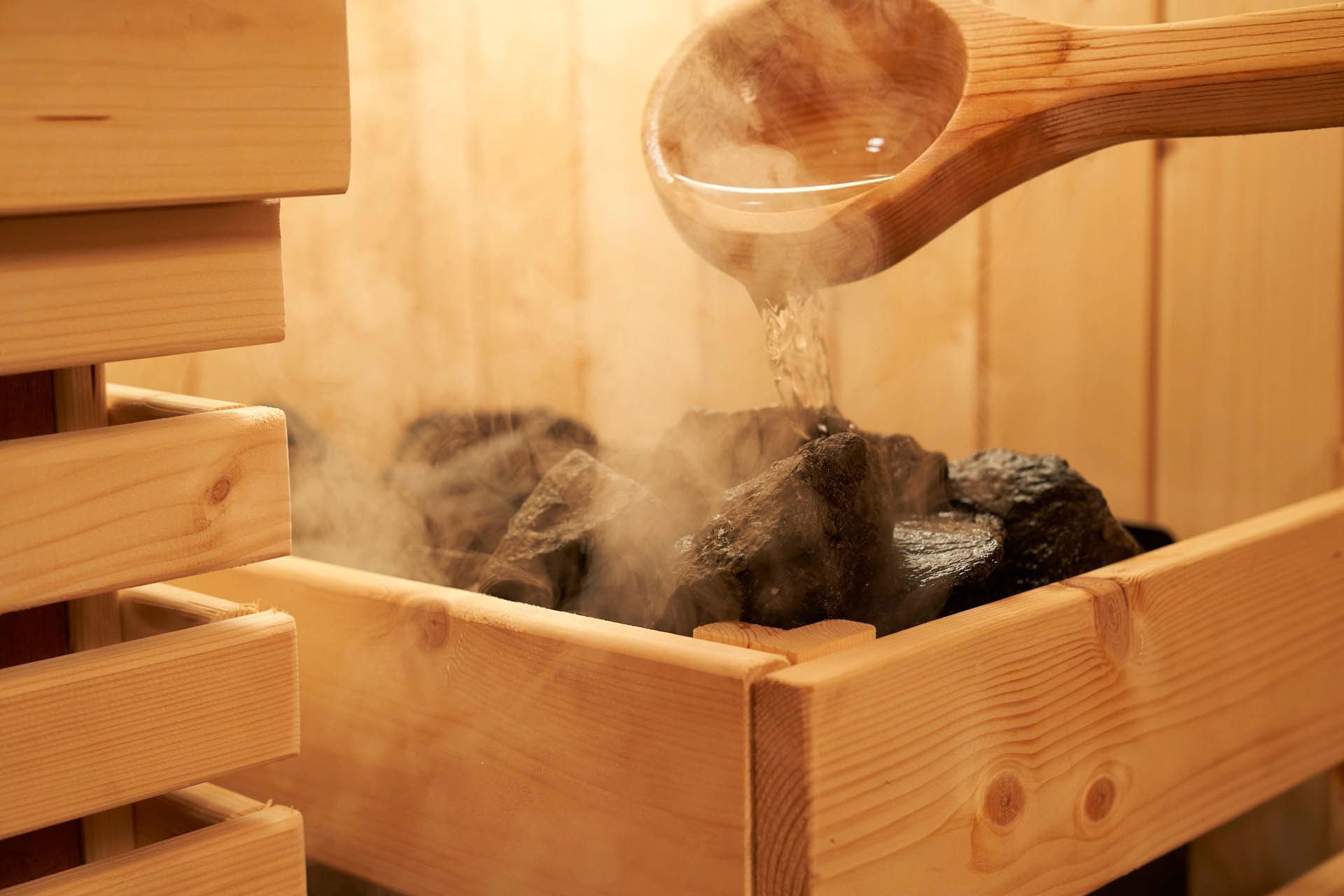 Image 5: A wooden ladle pours hot water over sauna stones, releasing steam in the warm masonry setting at Orchard Grand Court