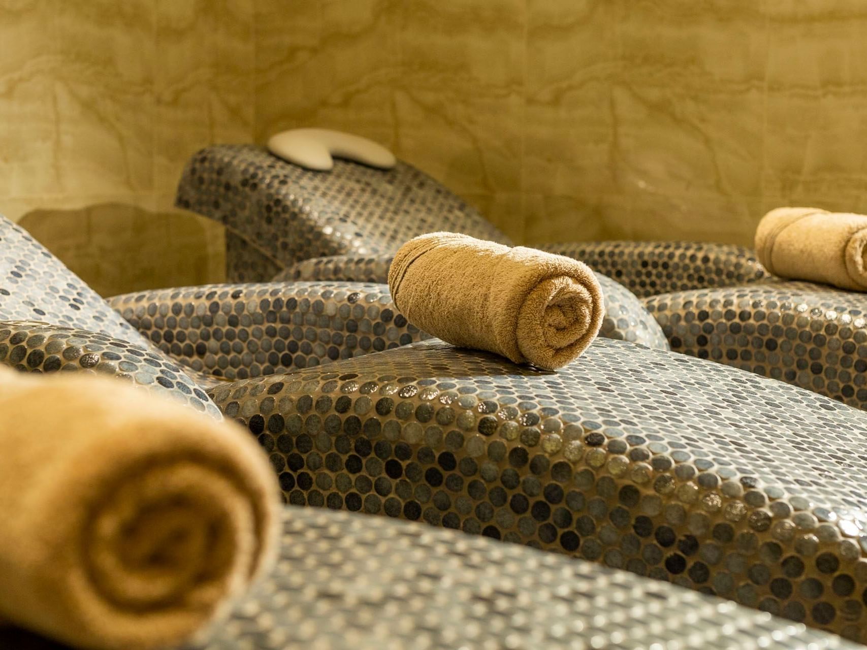 Four lounge chairs with rolled towels and a head pillow in a room with a beige wall.