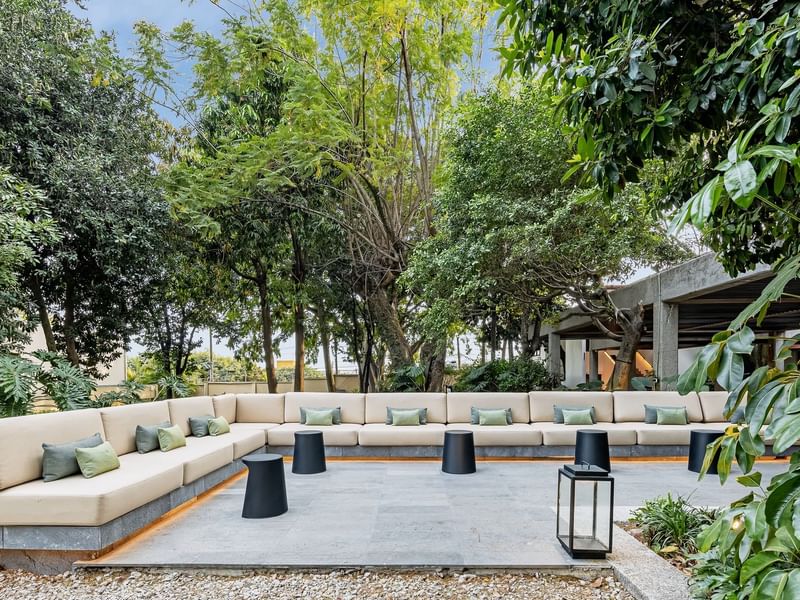 Outdoor seating area with large sofa, black stools, and lantern amid lush trees at Grand Fiesta Americana