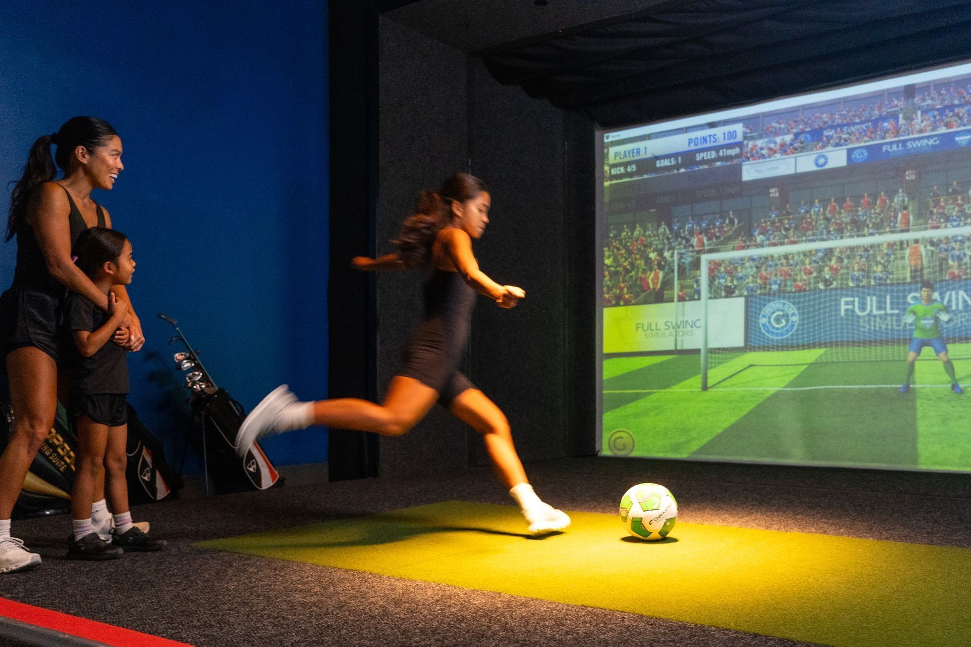 Girl playing soccer simulation game with soccer ball on mat, watched by a woman and a child.