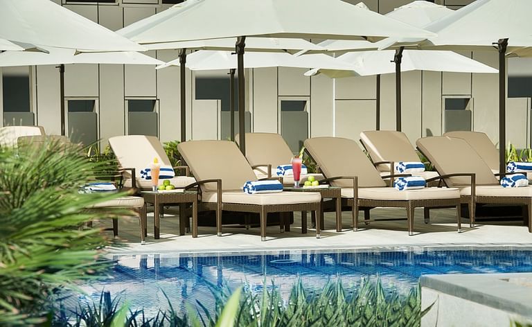 Poolside loungers and umbrellas on a sunny patio with beverages at DAMAC Maison Cour Jardin, a family hotel in Dubai